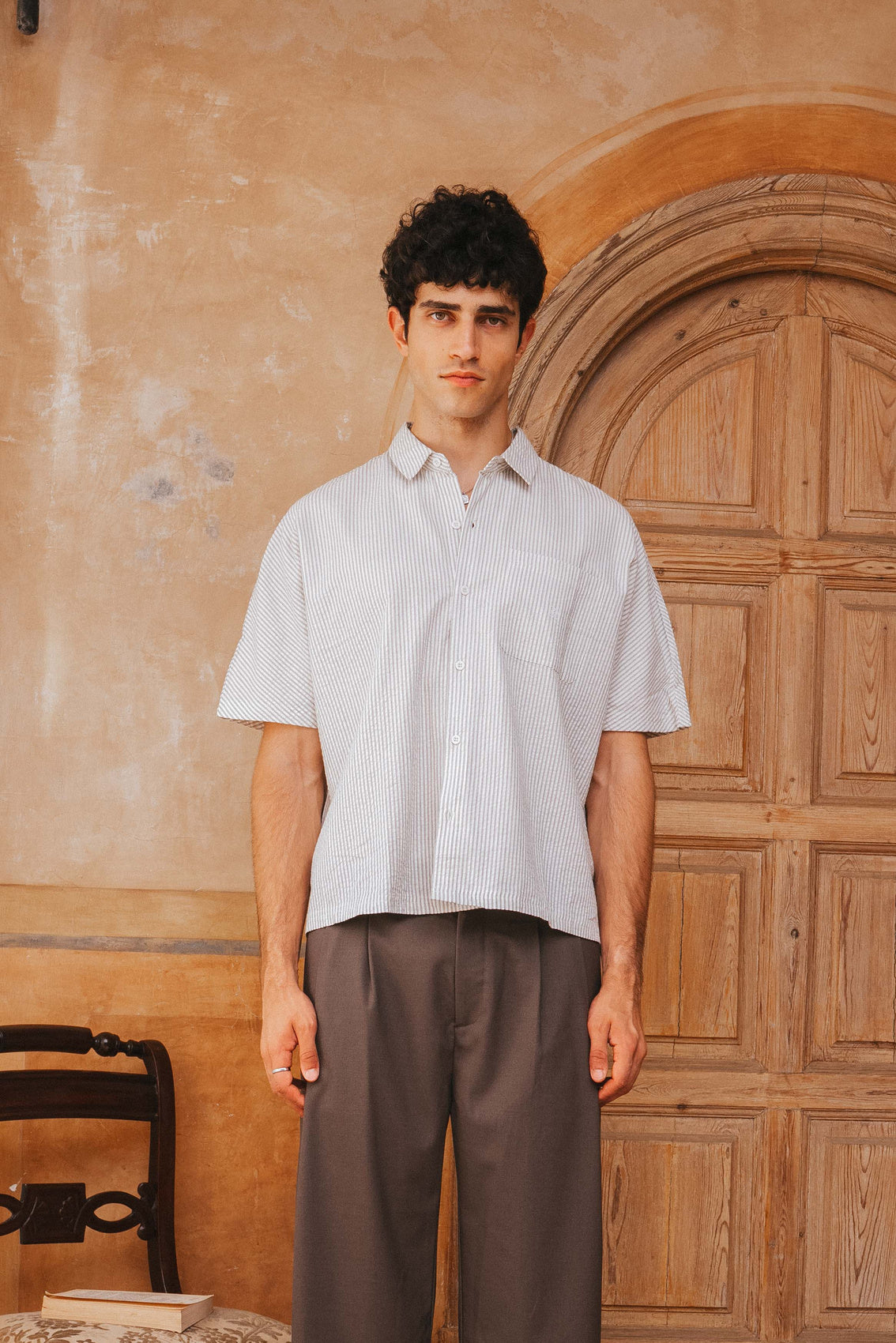 Man wearing a light-colored shirt and brown pants standing in front of a wooden door.