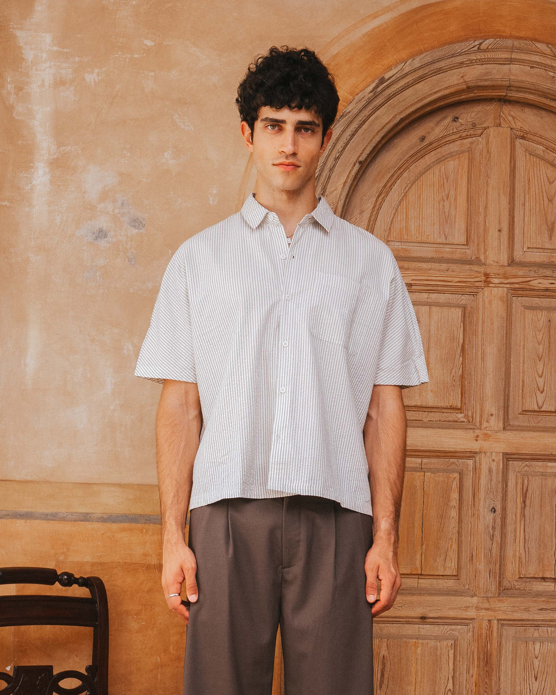 Man wearing a light-colored shirt and brown pants standing in front of a wooden door.