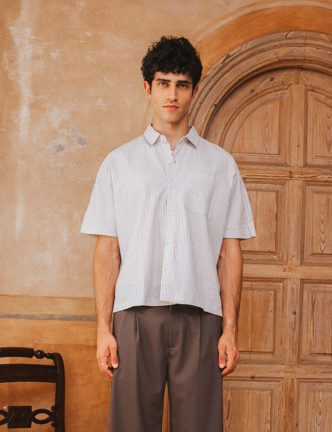 Man wearing a light-colored shirt and brown pants standing in front of a wooden door.