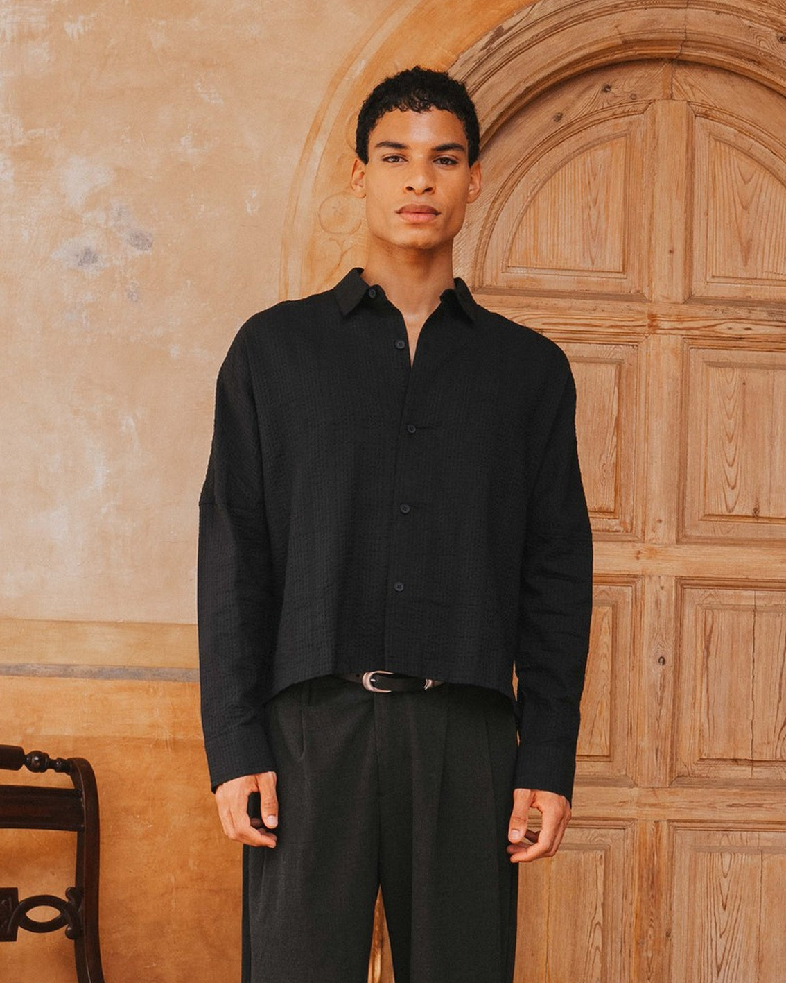 Man wearing a black shirt and pants standing in front of a wooden door.